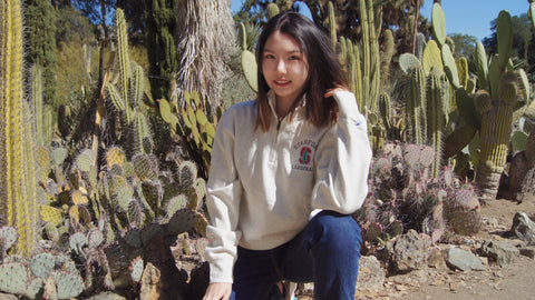 Crouching smiling student wearing Oatmeal colored quarter zip with Stanford "S" logo surrounded by white lettering reading "Stanford Cardinal"