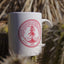 Close up image of a white ceramic mug with Stanford's shield printed in red letters. Background is out of focus cacti.