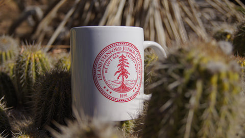 Close up image of a white ceramic mug with Stanford's shield printed in red letters. Background is out of focus cacti.